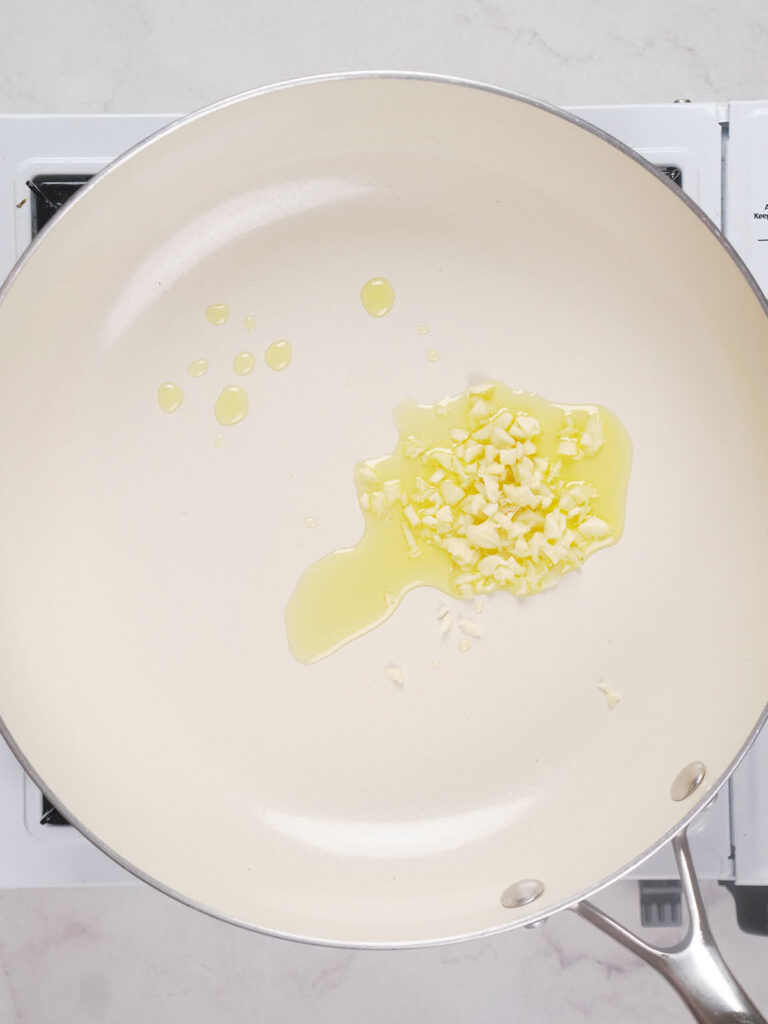 A light-colored nonstick frying pan on a stove with chopped garlic sautéing in oil.