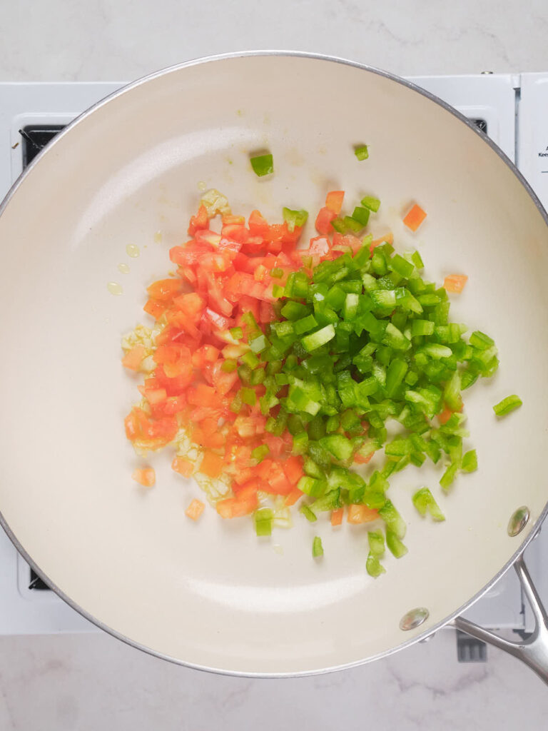 Chopped green bell peppers, tomatoes, and garlic cooking in a white skillet on a stovetop.