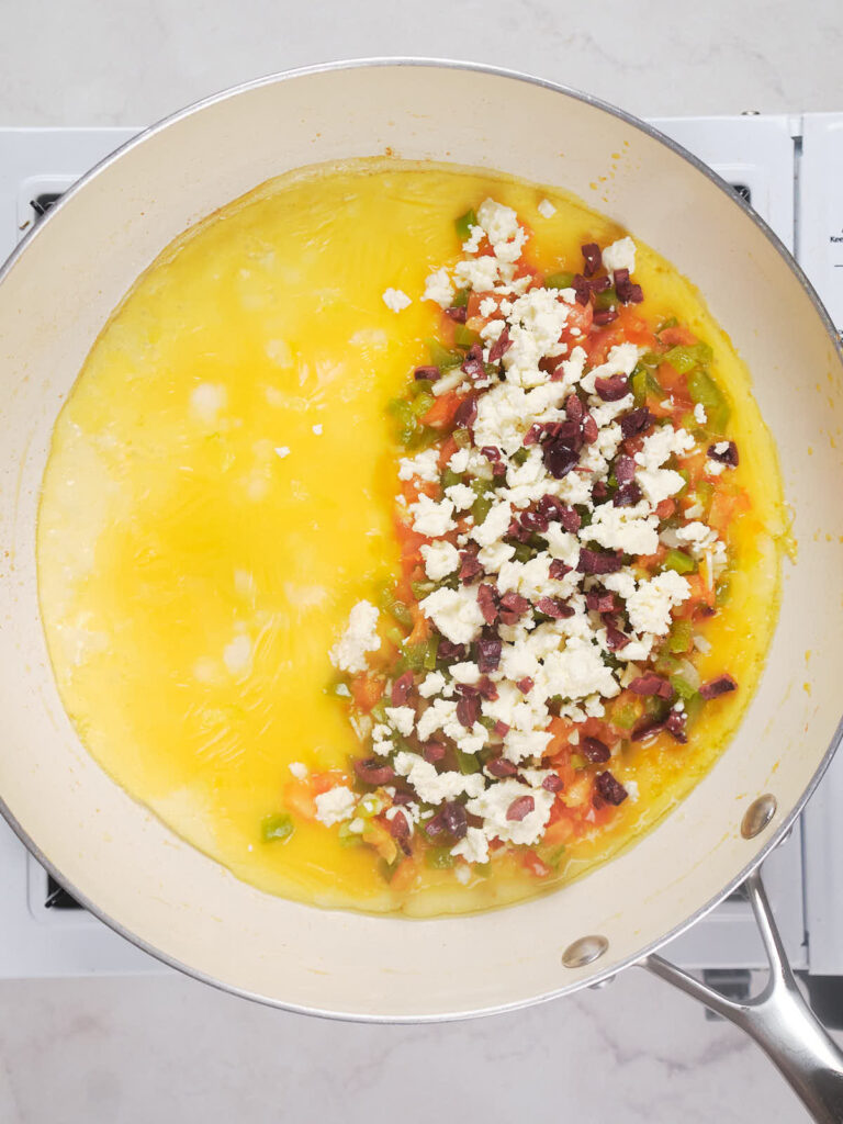 A half-cooked omelette in a pan, with one side filled with a mixture of diced vegetables, olives, and crumbled cheese.