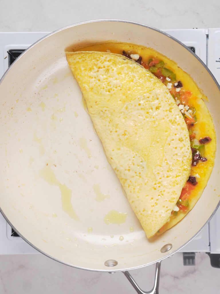 A partially folded omelet with visible pieces of vegetables and cheese cooking in a white skillet.
