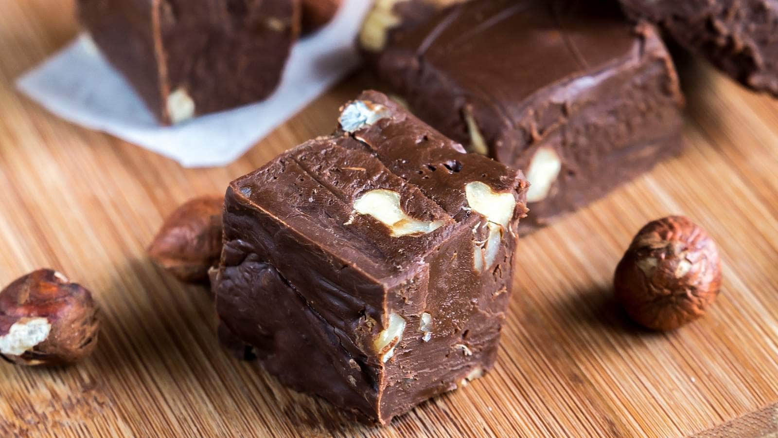 Close-up of squares of chocolate fudge with nut pieces on a wooden surface, accompanied by whole nuts.