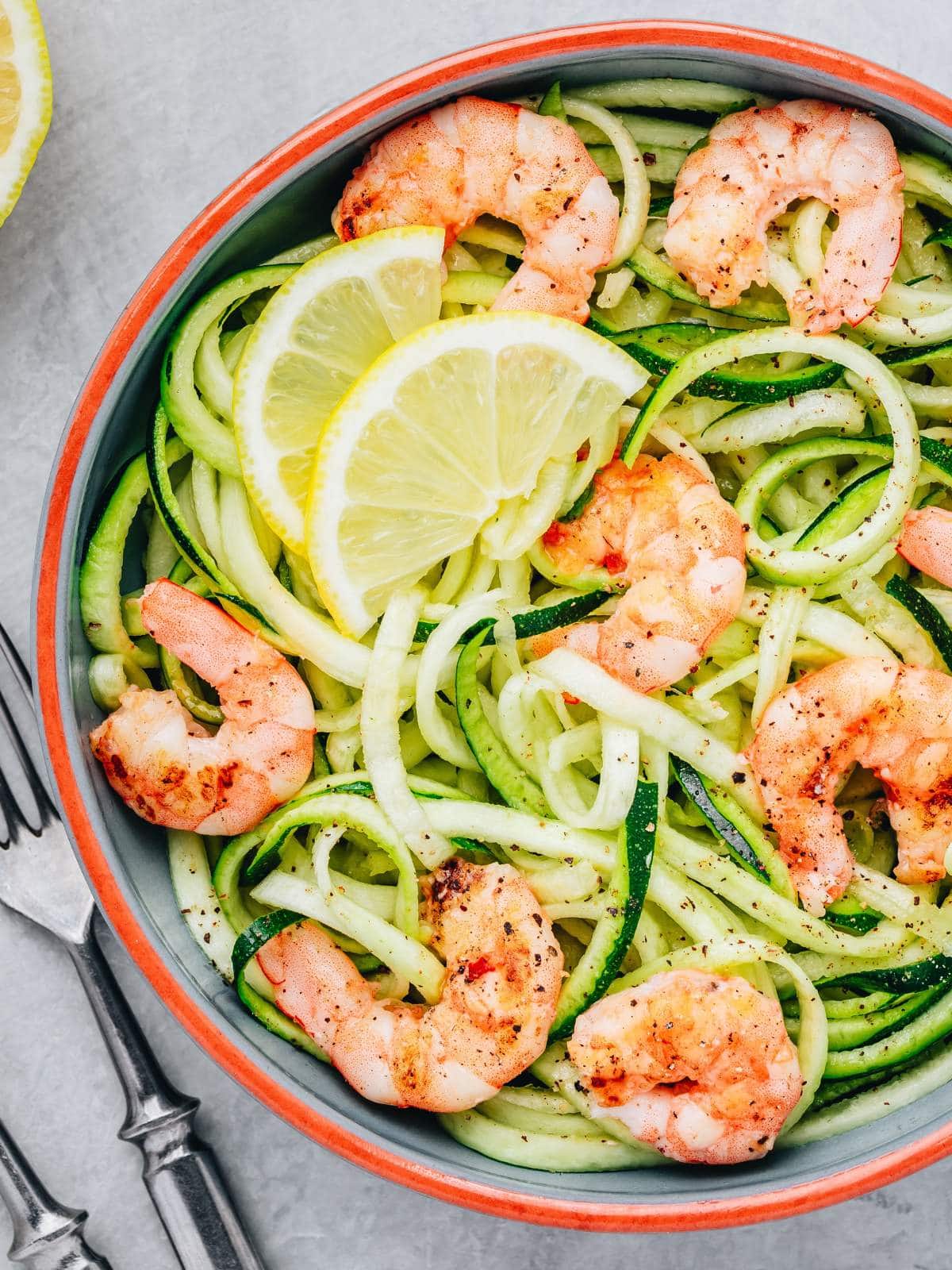 A bowl of zucchini noodles topped with grilled shrimp and lemon slices, with a fork and knife on the side.