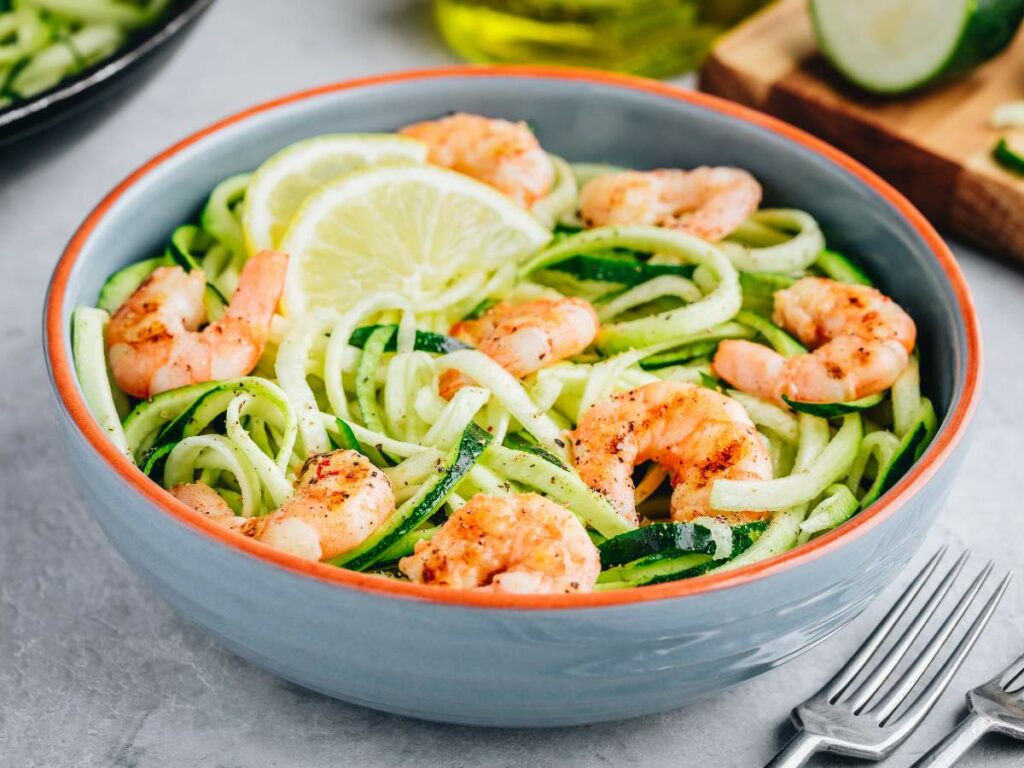 A blue bowl contains zucchini noodles with shrimp, garnished with lemon slices. There are a fork and knife placed nearby.