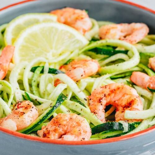 A bowl of zucchini noodles with cooked shrimp and lemon slices, garnished with black pepper, placed beside a fork.