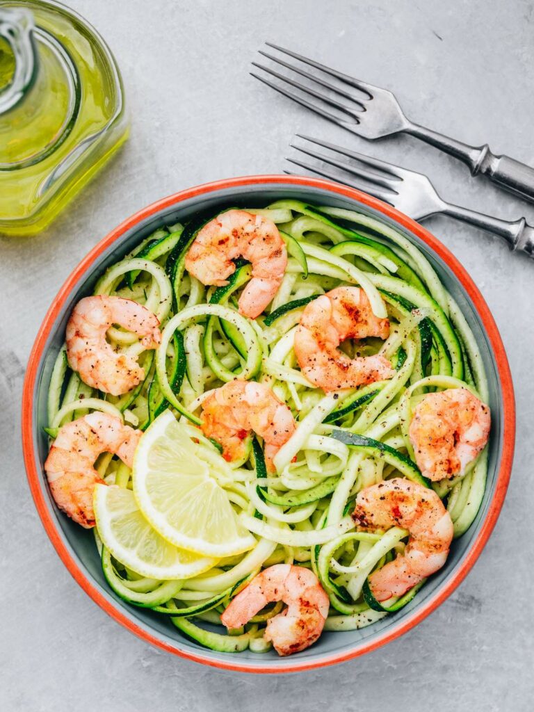 A bowl of zucchini noodles topped with cooked shrimp and lemon slices, with two forks and a bottle of olive oil next to it.