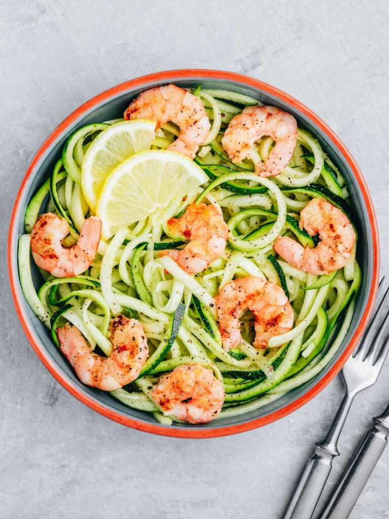 A bowl of zucchini noodles topped with shrimp and lemon slices. Two forks are placed next to the bowl on a gray surface.