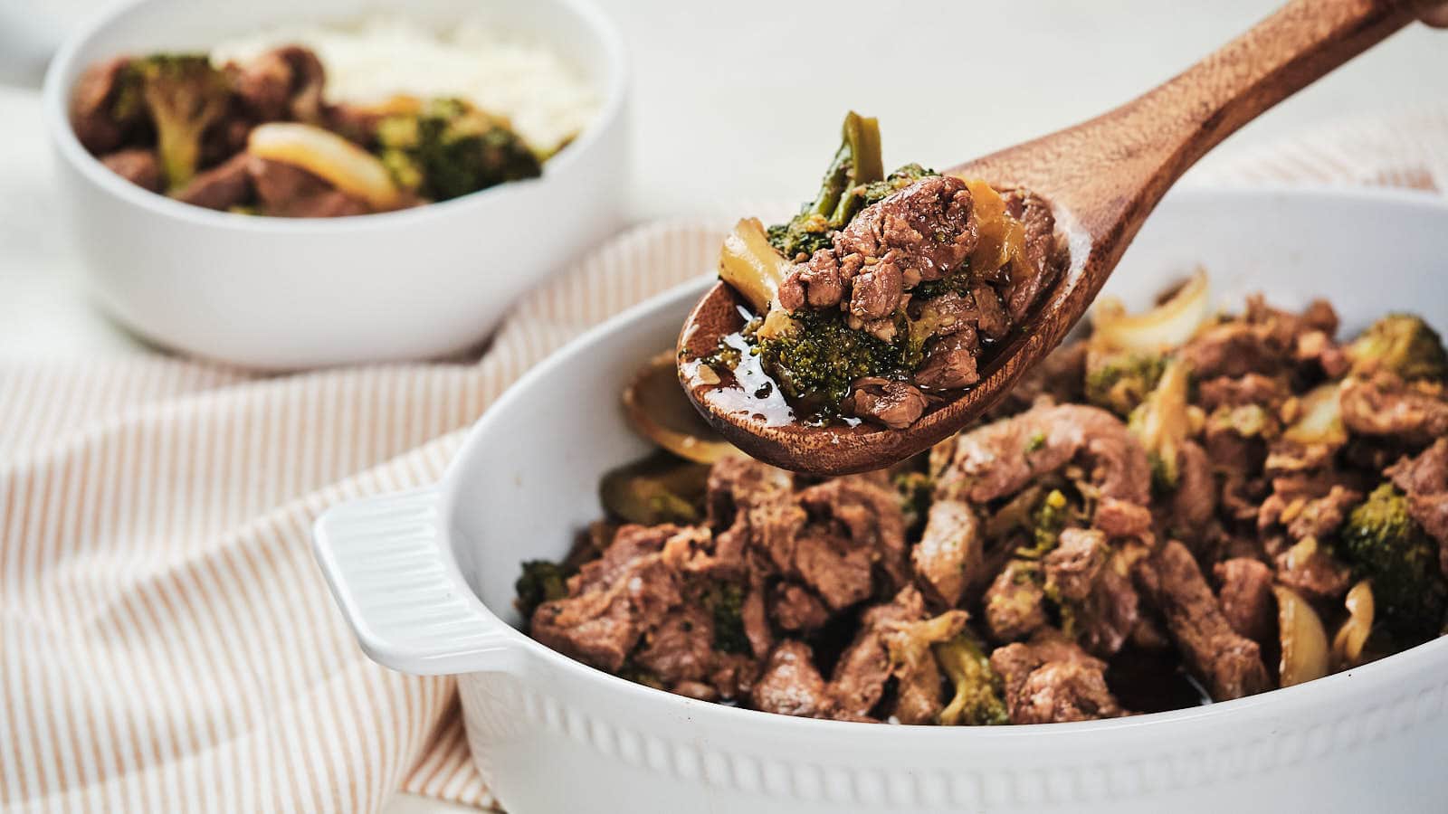 A wooden spoon serving beef and broccoli from a white baking dish, with a side of rice visible in the background.