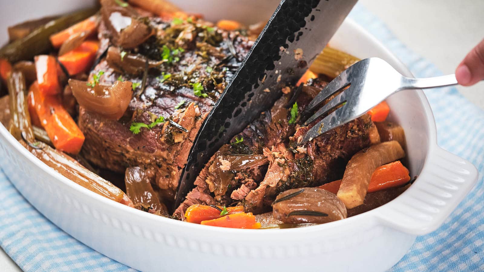 A person uses a fork to hold a cooked pot roast in a white baking dish while cutting it with a large knife. The dish contains cooked carrots, onions, and herbs.