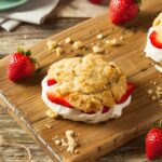 A wooden board holds a biscuit sandwich with whipped cream and strawberry slices.