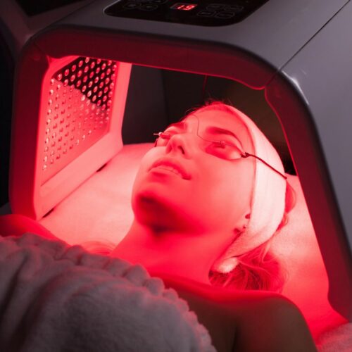 A person lying under a red LED light therapy device, wearing protective goggles and a headband.