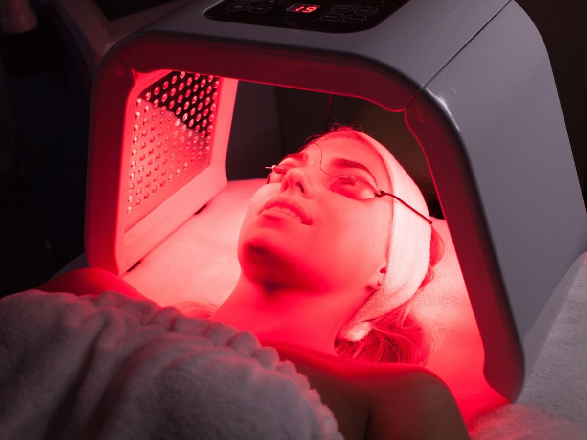 A person lying under a red LED light therapy device, wearing protective goggles and a headband.