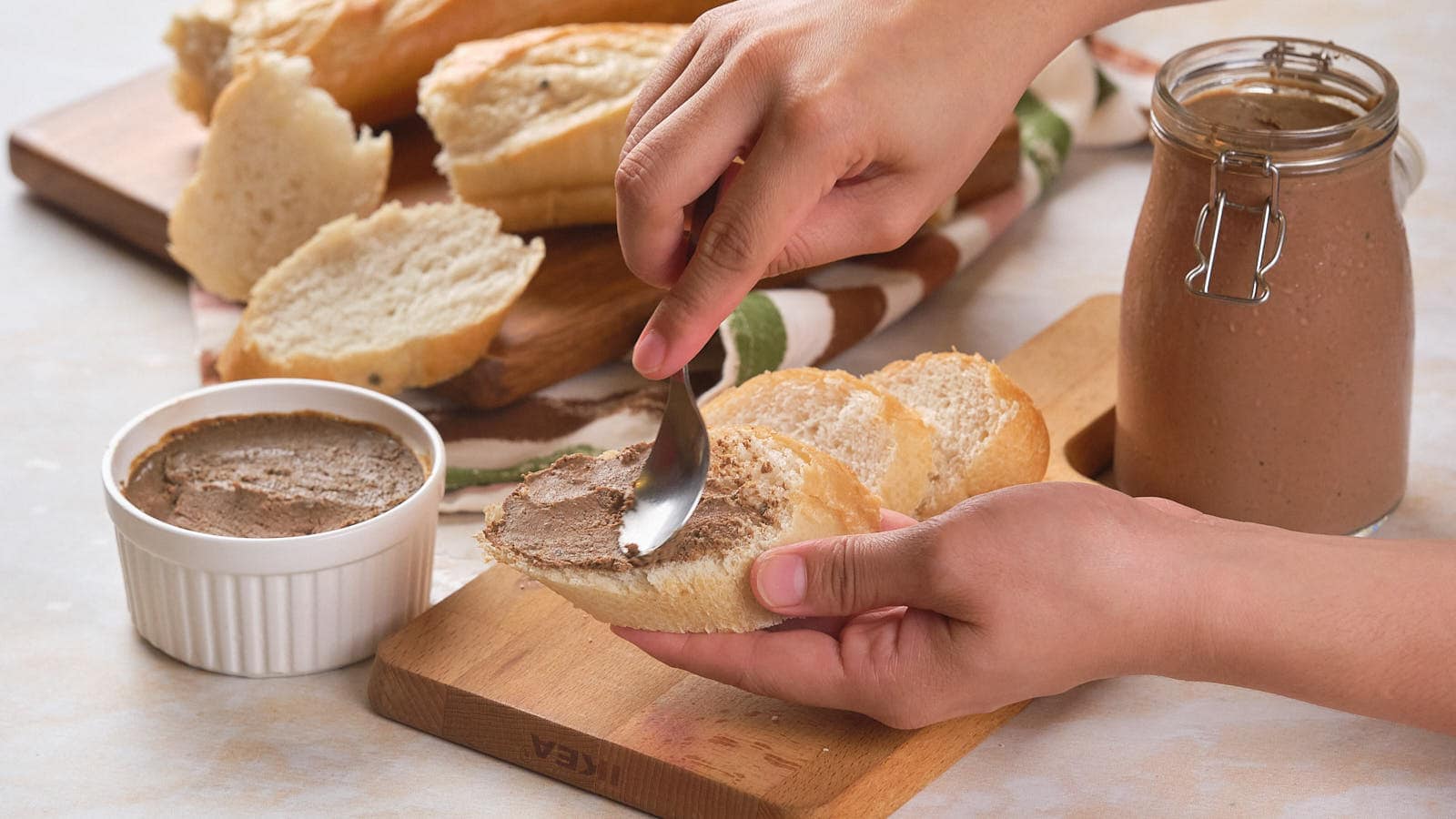 A person spreads beef pâté on a piece of bread. Several slices of bread and a jar of pâté are on the table.