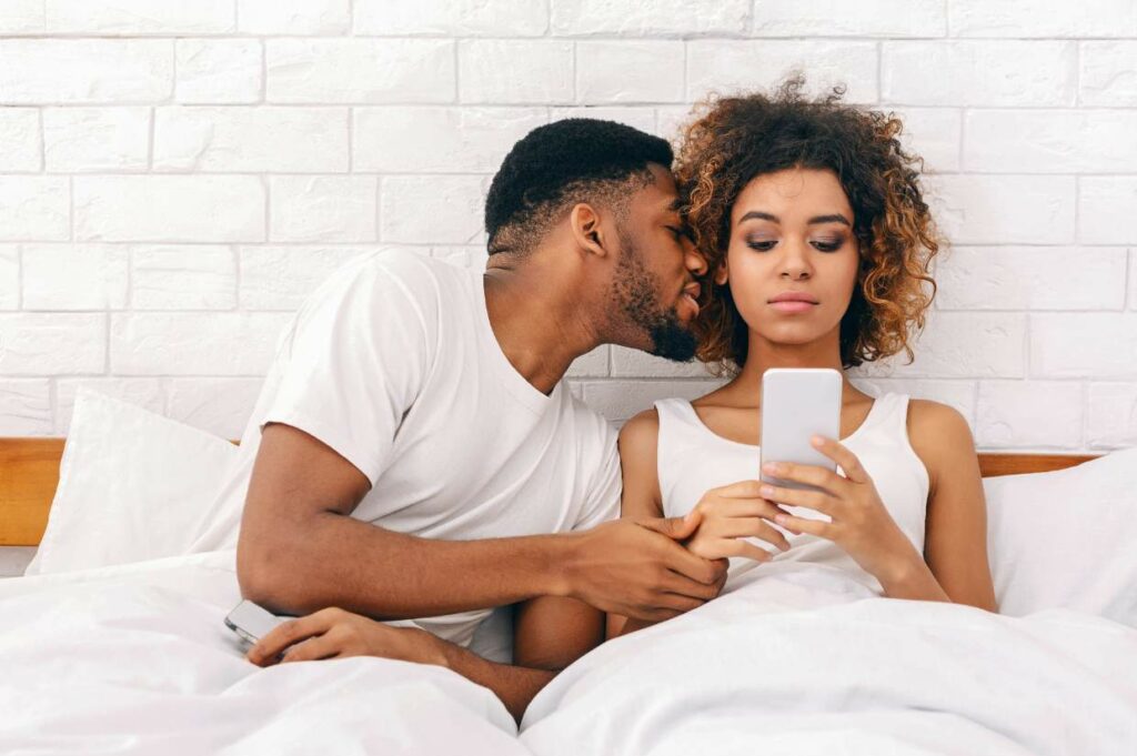 A man whispers to a woman who looks at her phone while lying side by side in bed wearing white clothes.