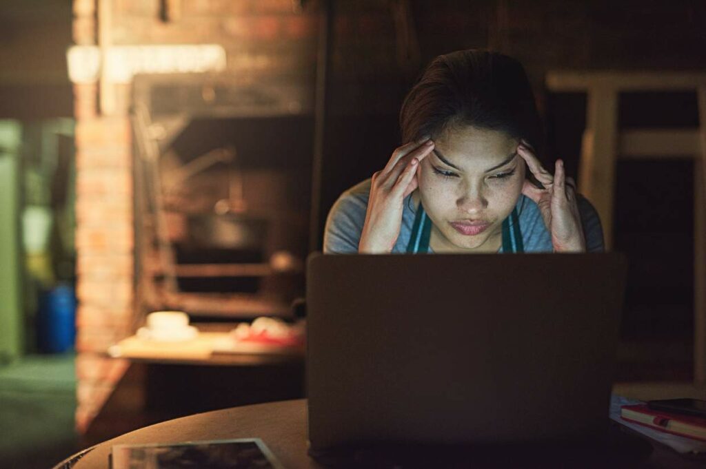 A person sits in front of a laptop, looking stressed with both hands touching their temples in a dimly lit room.