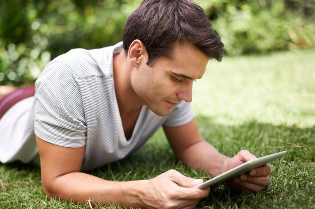 A man lies on the grass, looking at a tablet in his hands.