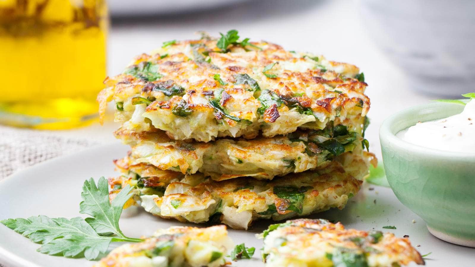 Three cabbage fritters stacked on a plate with a dollop of sour cream on the right and a sprig of parsley on the left.