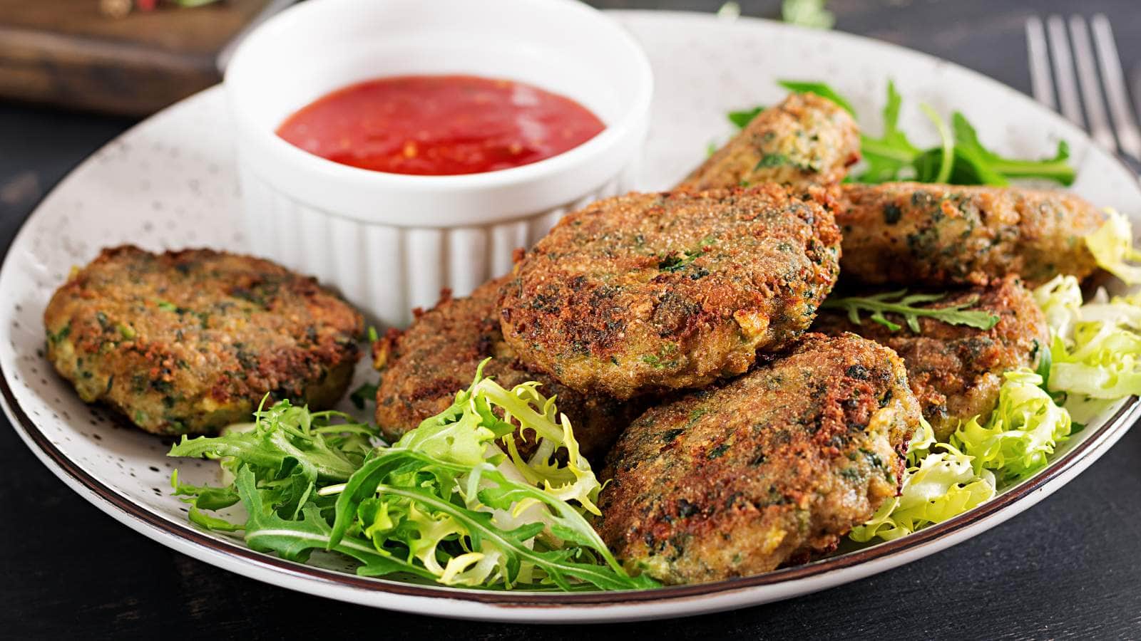 A plate of vegetable fritters on a bed of greens, served with a side of red dipping sauce in a small white bowl.