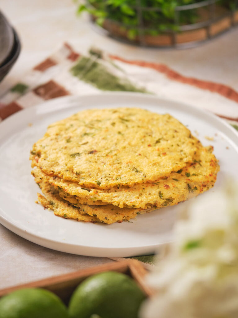 A stack of four herb-infused cauliflower tortilla is placed on a white plate in a kitchen with a blurred background.