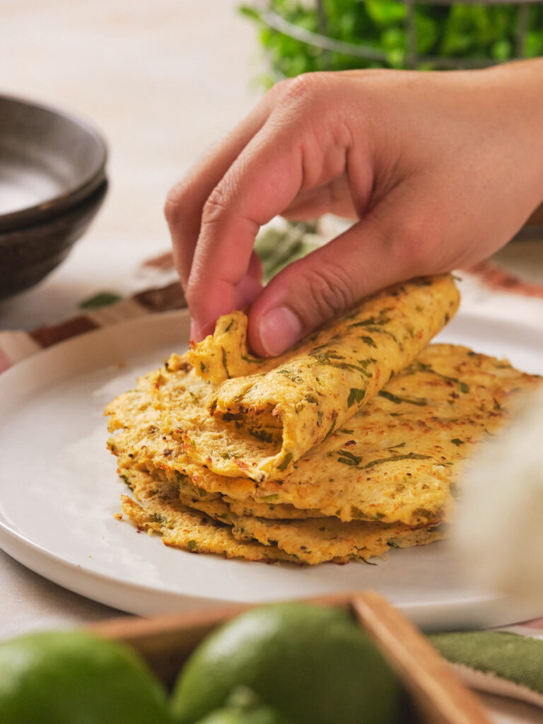 A hand lifting a piece of cauliflower tortilla from a stack on a white plate.