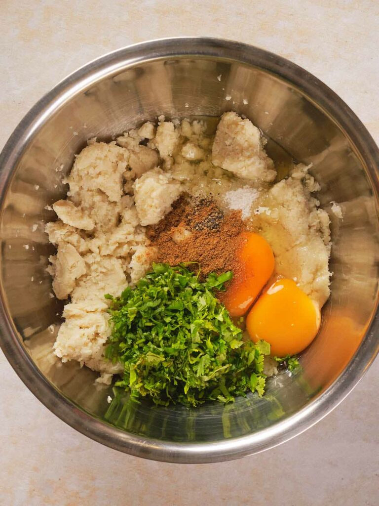 A metal bowl containing mashed cauliflower, two raw eggs, chopped herbs, and various seasonings on a light-colored surface.