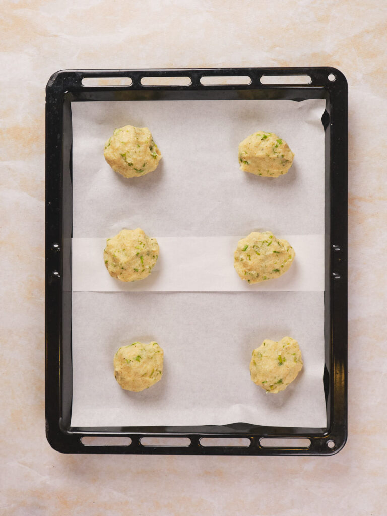 A baking tray holds six evenly spaced cauliflower tortilla balls on parchment paper, ready for baking.