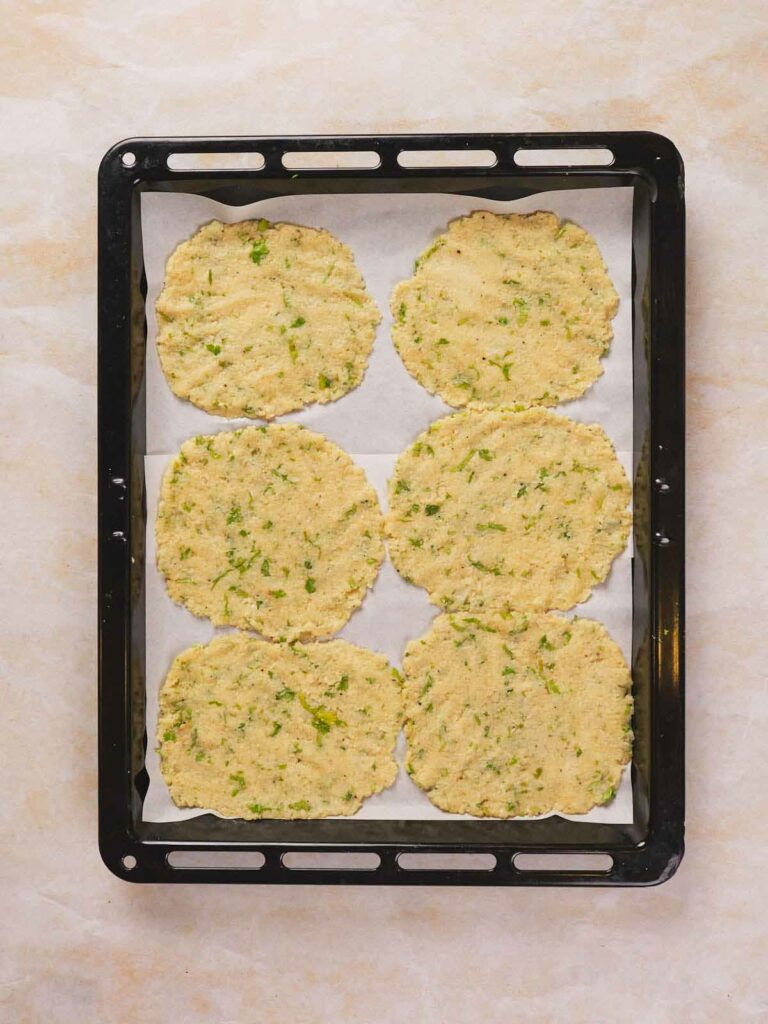 A baking tray with six uncooked cauliflower tortilla with visible green herbs.