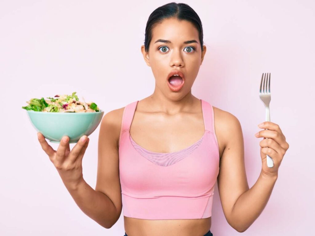 A person wearing a pink sports bra holds a bowl of salad in one hand and a fork in the other.