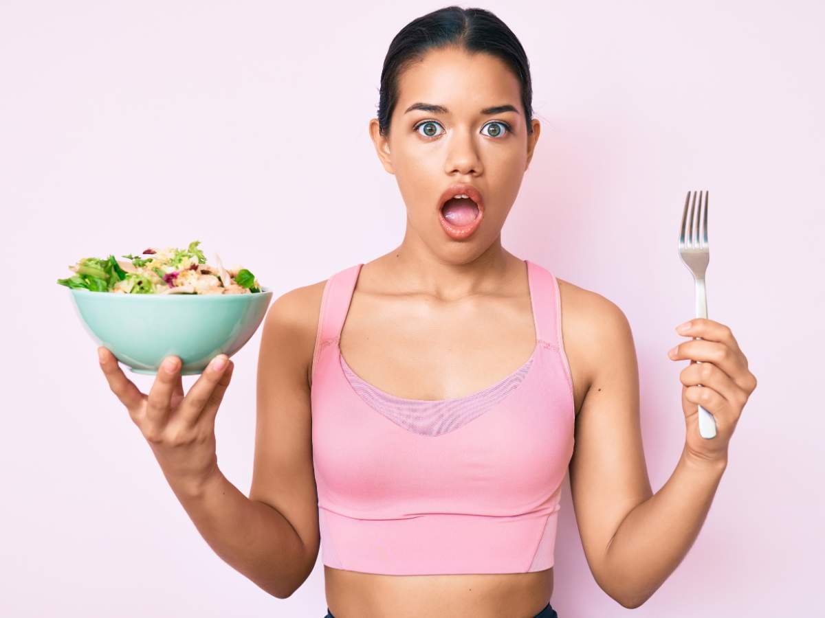 A person wearing a pink sports bra holds a bowl of salad in one hand and a fork in the other.