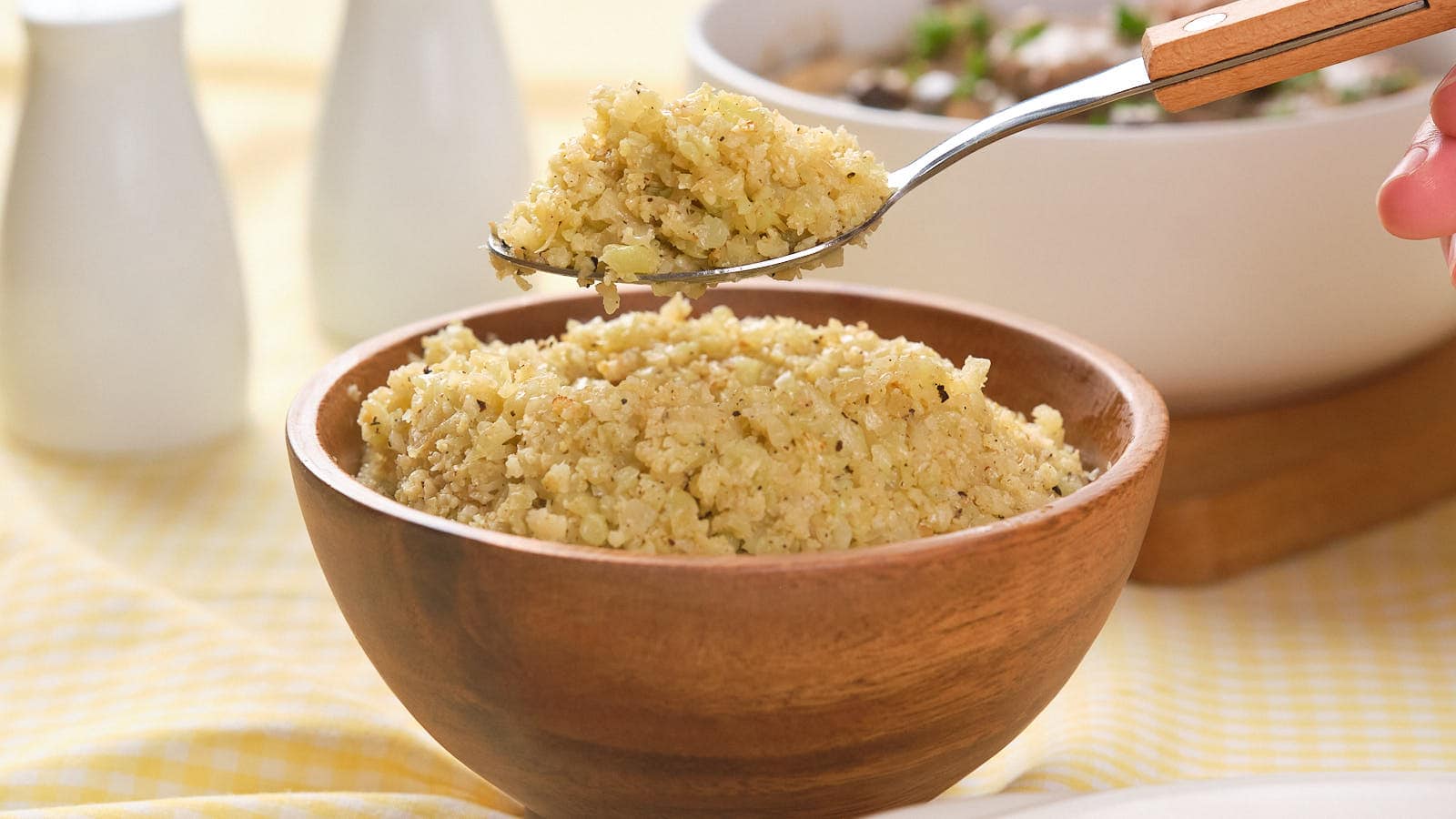 A spoonful of cooked cauliflower rice hovers over a wooden bowl full of the same dish, with additional bowls in the background.