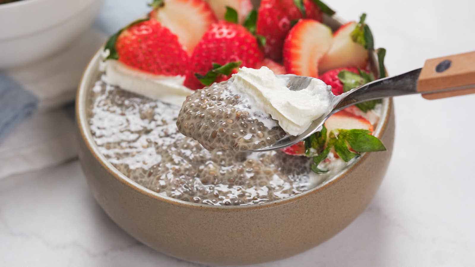 A bowl of chia pudding topped with sliced strawberries and a dollop of whipped cream, with a spoon lifting a portion.