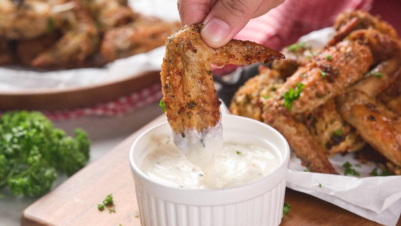 A hand dips a parmesan chicken wing into a small ramekin filled with white dipping sauce.