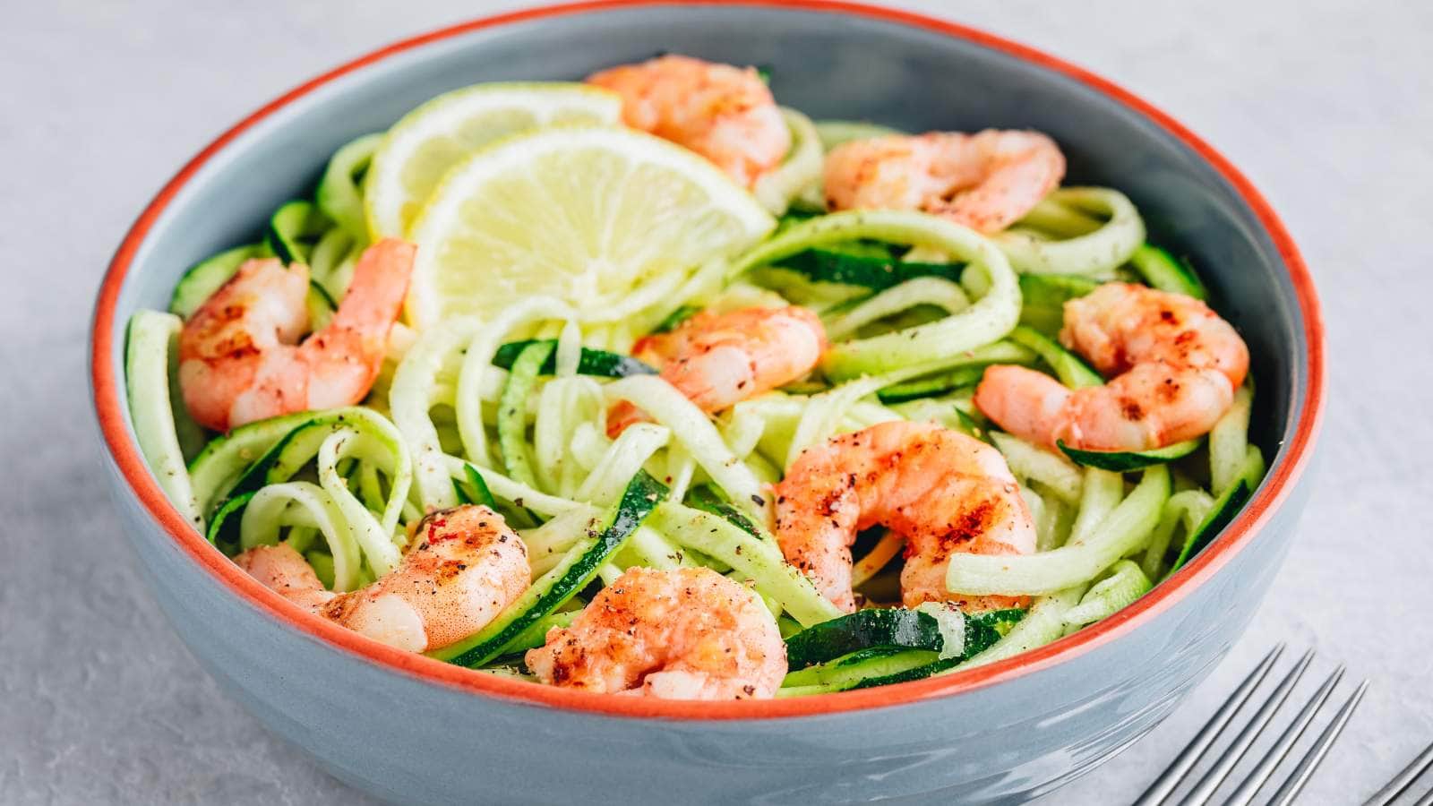 A bowl of shrimp and zucchini noodles garnished with lemon slices, placed on a light grey surface next to a fork.