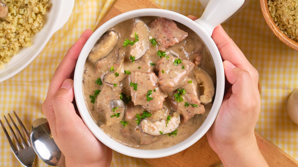 A person holds a bowl of creamy beef stroganoff garnished with parsley.