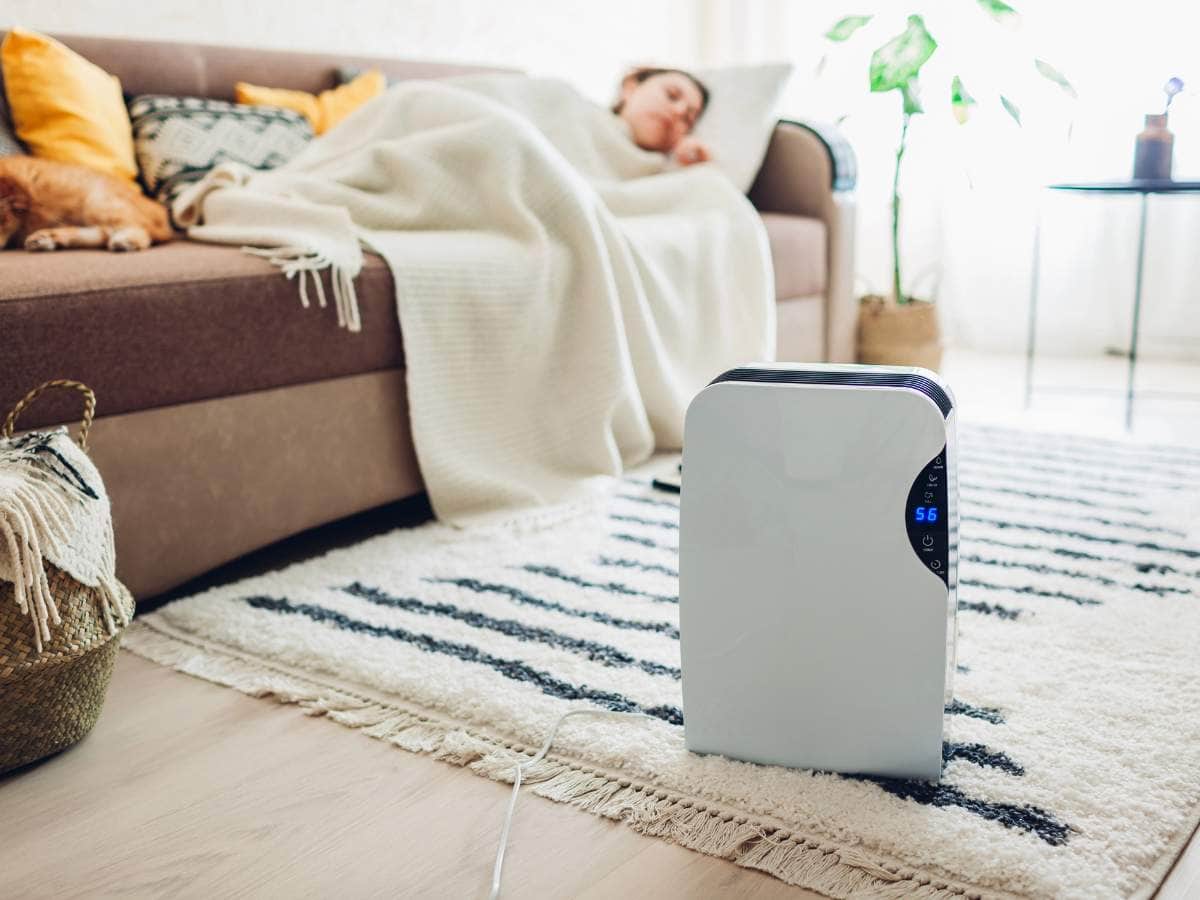 A person resting on a couch covered with a blanket with a white dehumidifier running on a striped rug on the floor.