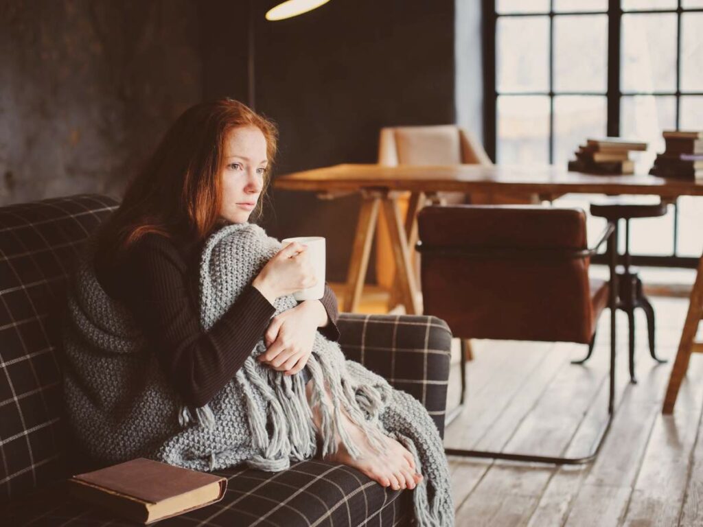 A person with red hair sits on a couch, wrapped in a blanket and holding a mug.