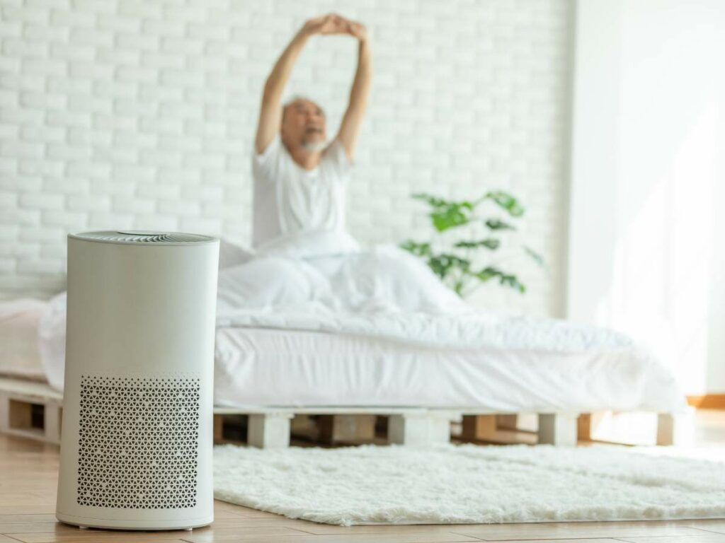 A white air purifier in a bedroom, with a person stretching on a bed in the background.