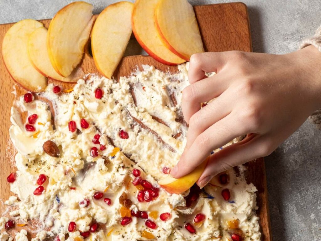 Hand dipping apple slice into a creamy spread topped with pomegranate seeds, nuts, and herbs on a wooden board.