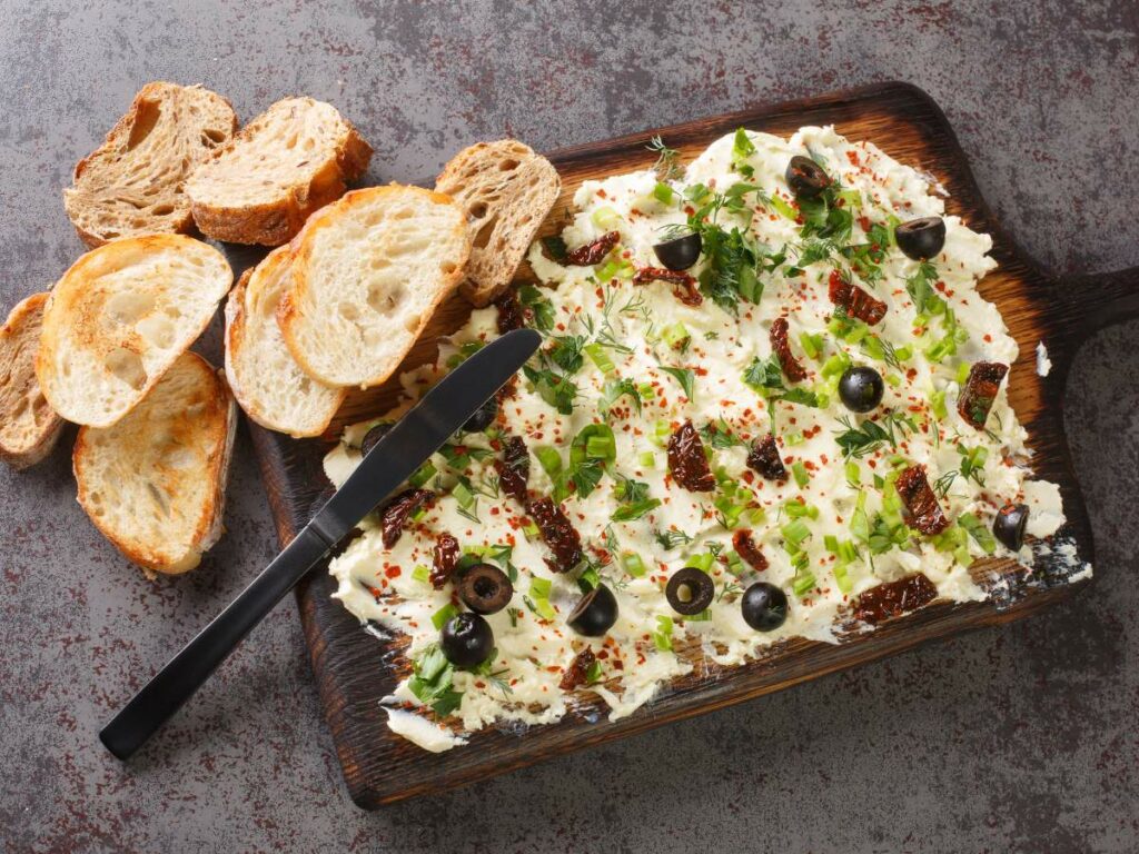 A wooden platter with a spread of butter topped with olives, herbs, and seasoning, accompanied by sliced bread.