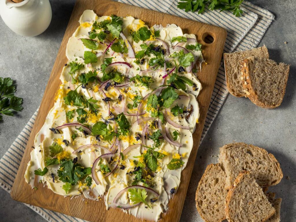 A wooden board holds a spread, garnished with red onion slices, fresh herbs, and lemon zest.