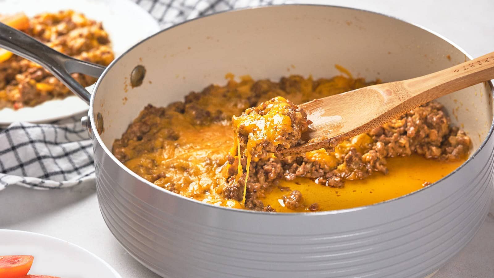 A wooden spoon lifts a serving of cheeseburger-skillet, with a plate of the same food visible in the background.