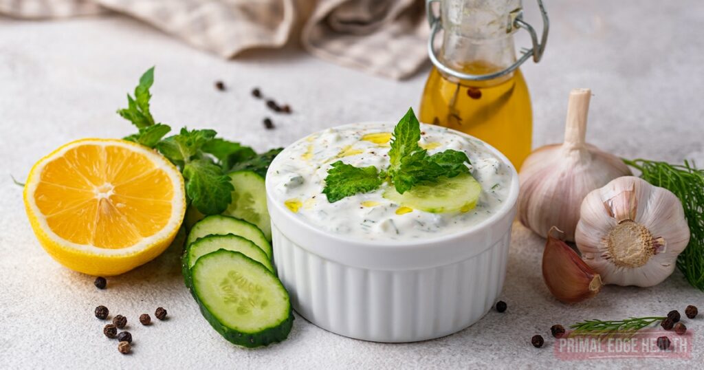 A bowl of tzatziki sauce garnished with mint, surrounded by lemon, cucumber slices, garlic, and a bottle of olive oil on a light surface.