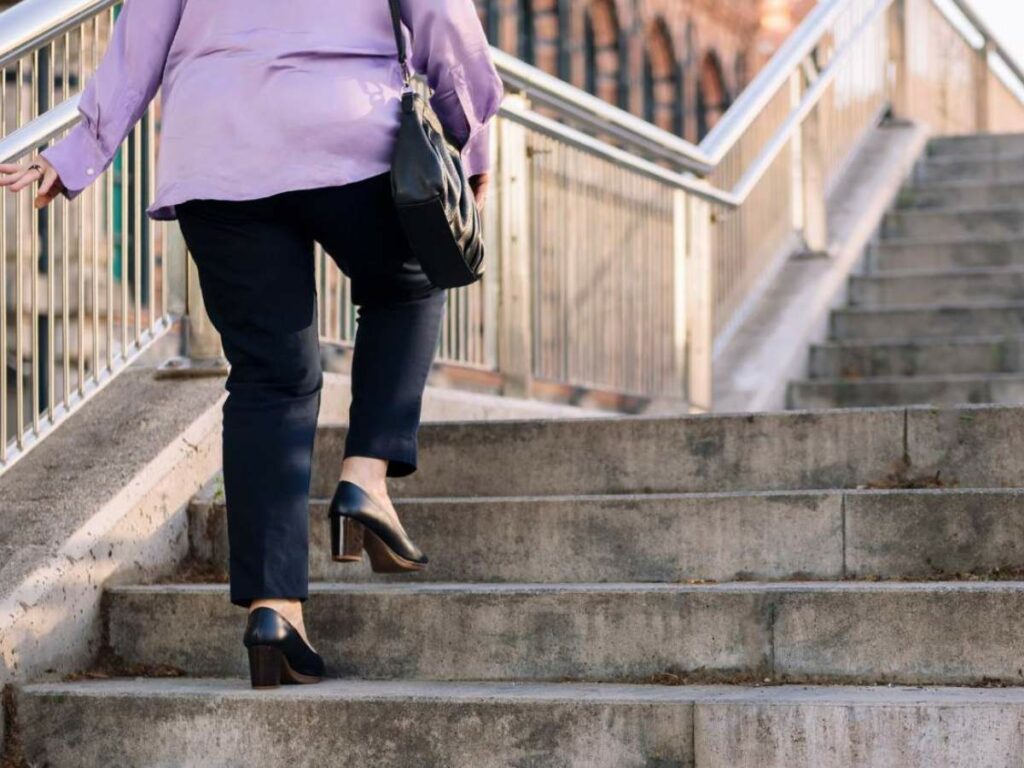 A person wearing a light purple shirt, dark pants, and black heels, is walking up a concrete staircase with metal railings.