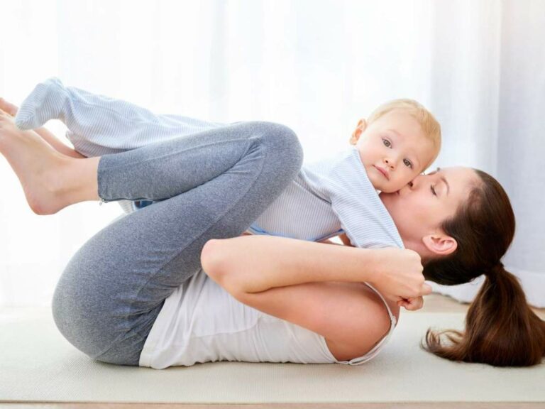 A woman in athletic wear lies on her back and lifts a baby in her arms and legs while performing an exercise on a yoga mat.