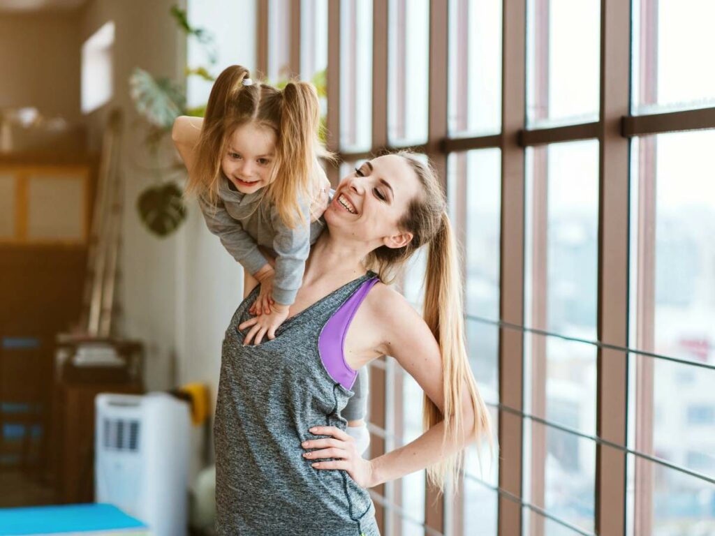 A woman in athletic wear smiles while holding a young girl with pigtails on her shoulders.