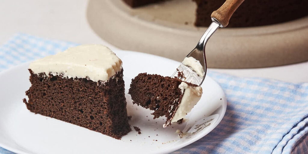 A slice of chocolate cake with white icing on a plate, with a fork lifting a piece.