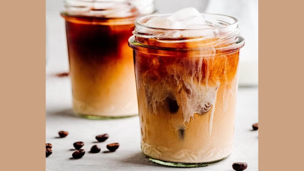 Two glass jars filled with iced coffee, with a gradient from light milk to dark coffee. Ice cubes and coffee beans are scattered around the jars on a light surface.