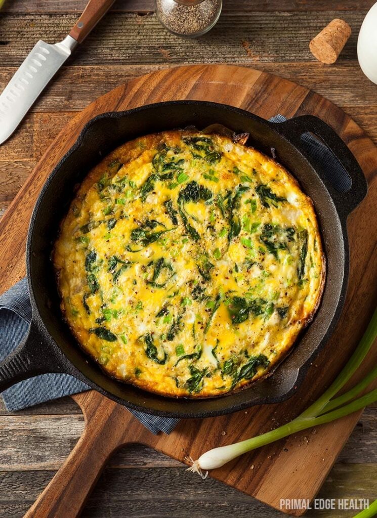 A frittata with greens, baked in a cast-iron skillet, is placed on a wooden cutting board next to green onions, a knife, and an egg—capturing the essence of primal edge health.