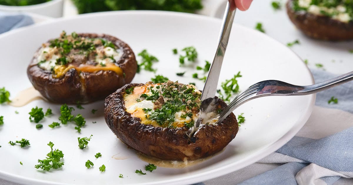 A person cuts into egg in mushroom with herbs and cheese on a white plate.