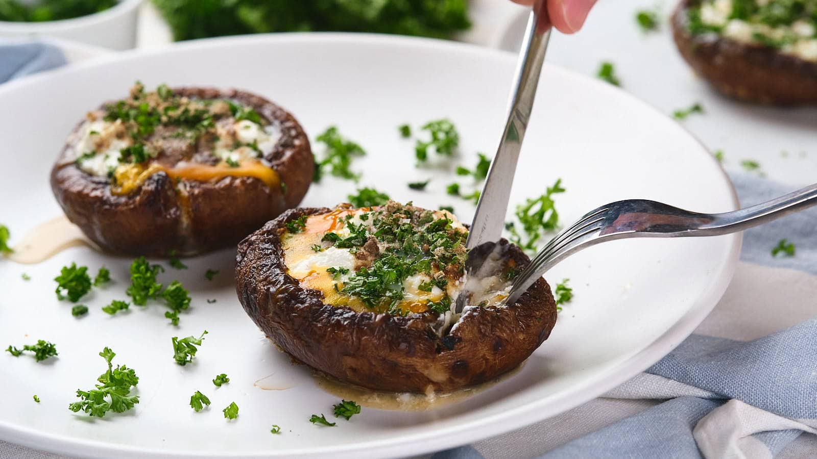 A fork and knife cutting into an egg in mushroom with green garnish visible in the background.