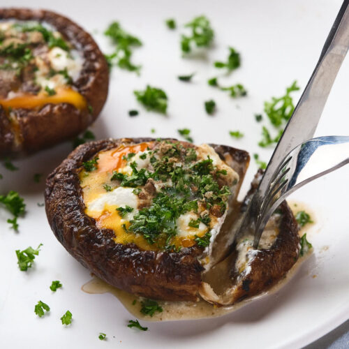 A fork slices into an egg in mushroom garnished with chopped parsley on a white plate.